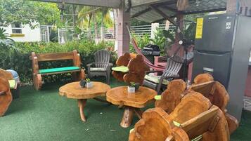A covered outdoor seating area with wooden furniture, a grill, and a refrigerator.