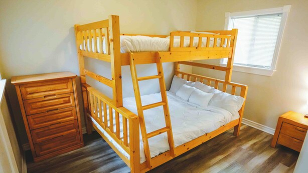 Ein Stockbett mit Holzleiter, eine hölzerne Kommode und ein kleiner Nachttisch in einem Zimmer mit Holzboden und einem Fenster mit Jalousien.