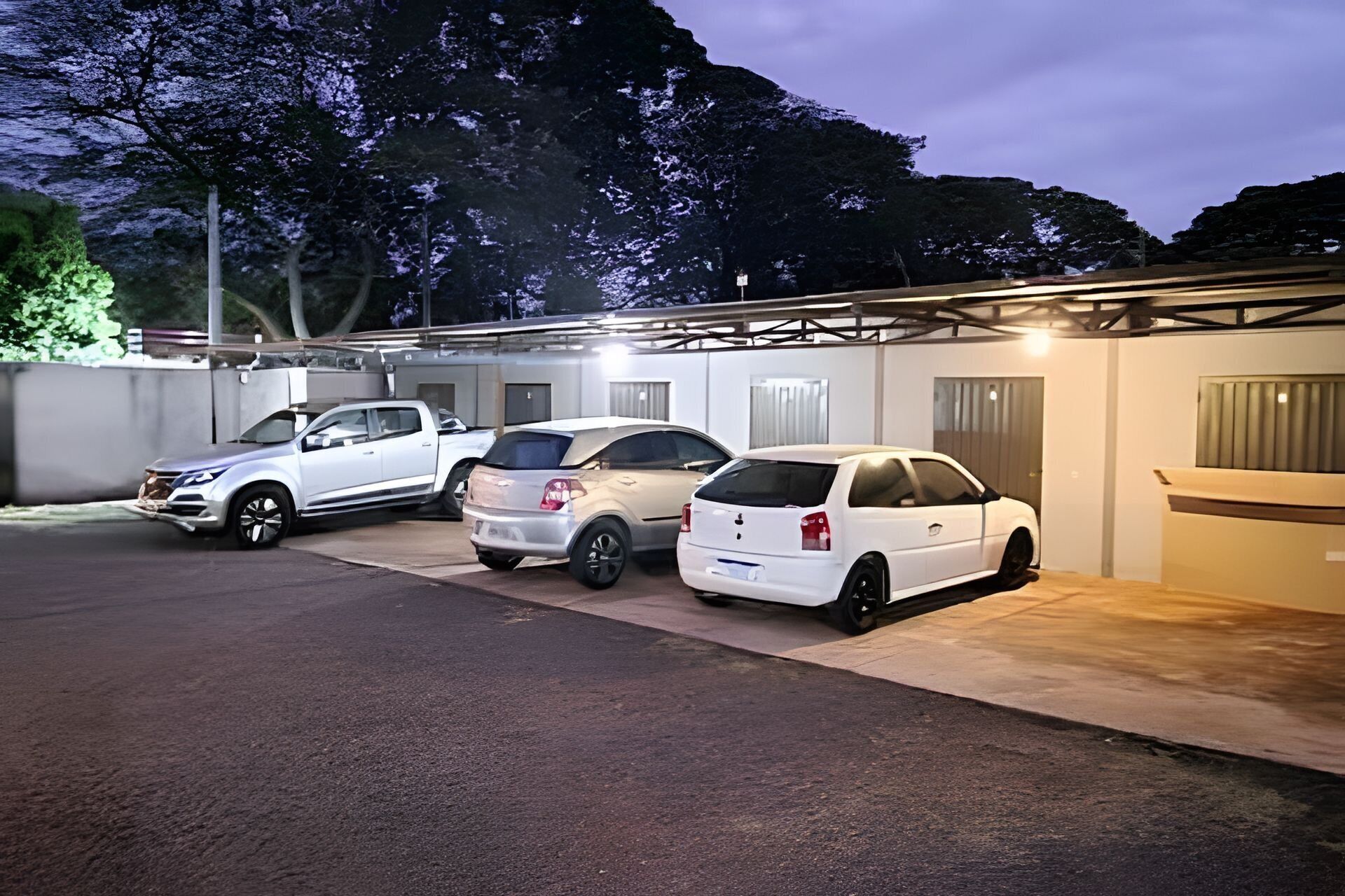 A parking area with three cars parked in a row, a covered structure with a roof, and a building with a door and windows.