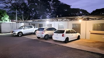 A parking area with three cars parked in a row, a covered structure with a roof, and a building with a door and windows.
