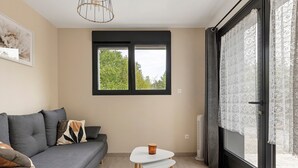 A living room with a grey sofa, a small white table, and a window with a view of trees.