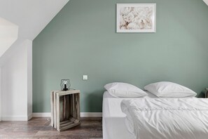 A bedroom with a sloped ceiling, a bed with white bedding, a small side table with a light, and a wall-mounted light switch.