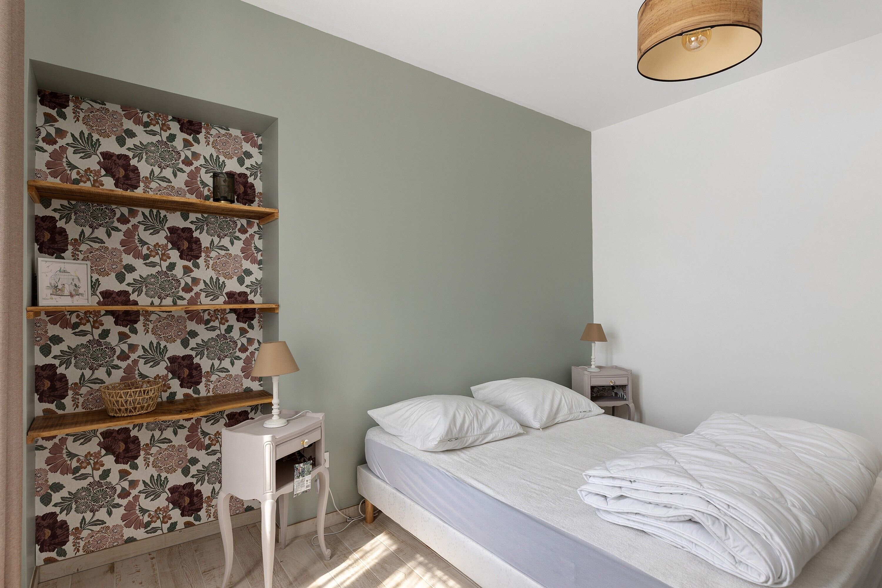A bedroom with a bed, a nightstand, a lamp, and a shelf with a floral wallpaper.