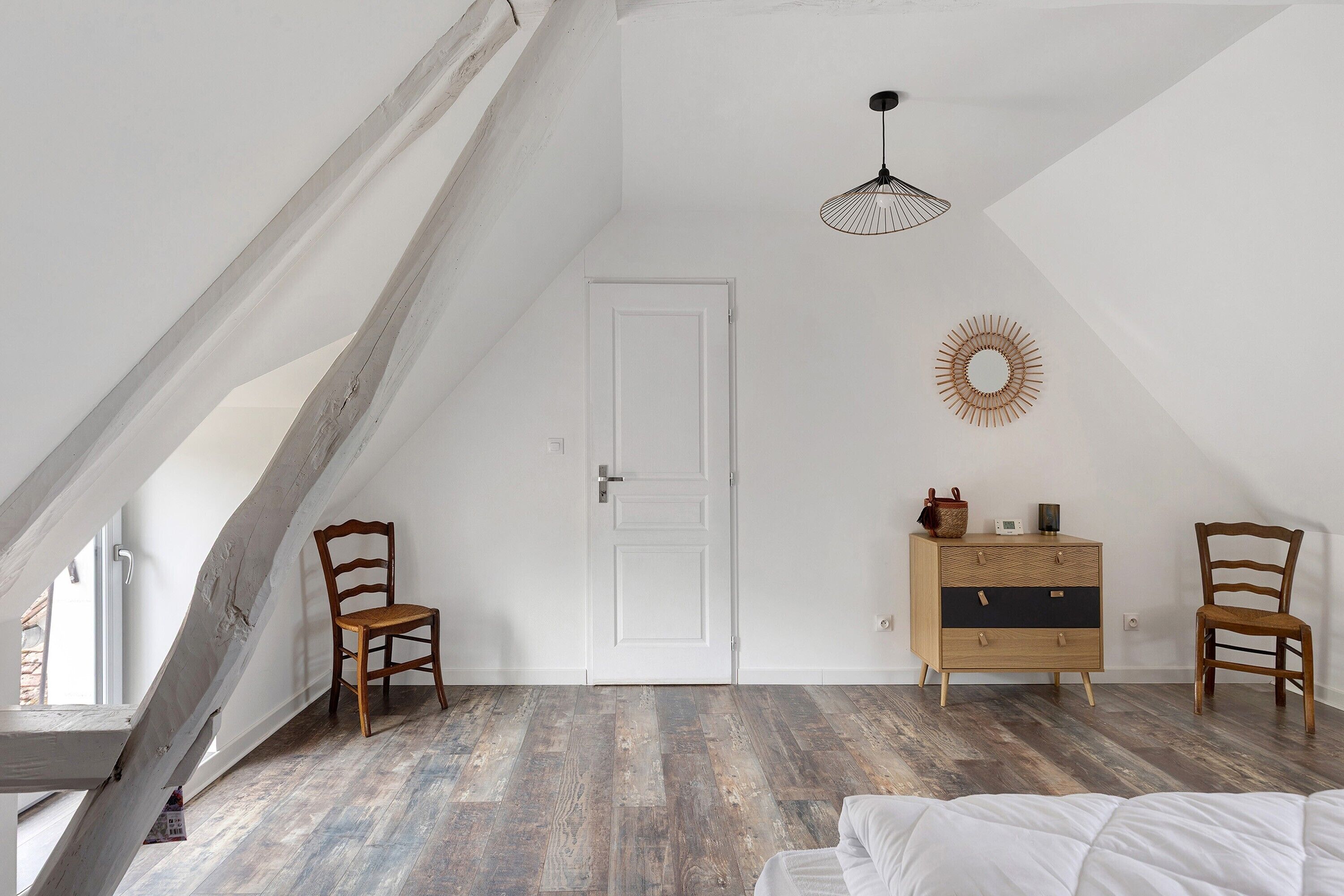 Une pièce avec un plafond en pente, une porte blanche, deux chaises en bois, une commode en bois, un luminaire suspendu et un objet décoratif fixé au mur.