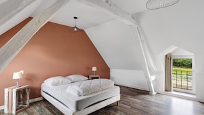 A bedroom with a sloped ceiling, a bed, a nightstand, a lamp, and a view of greenery through a window.