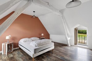 A bedroom with a sloped ceiling, a bed, a nightstand, a lamp, and a view of greenery through a window.
