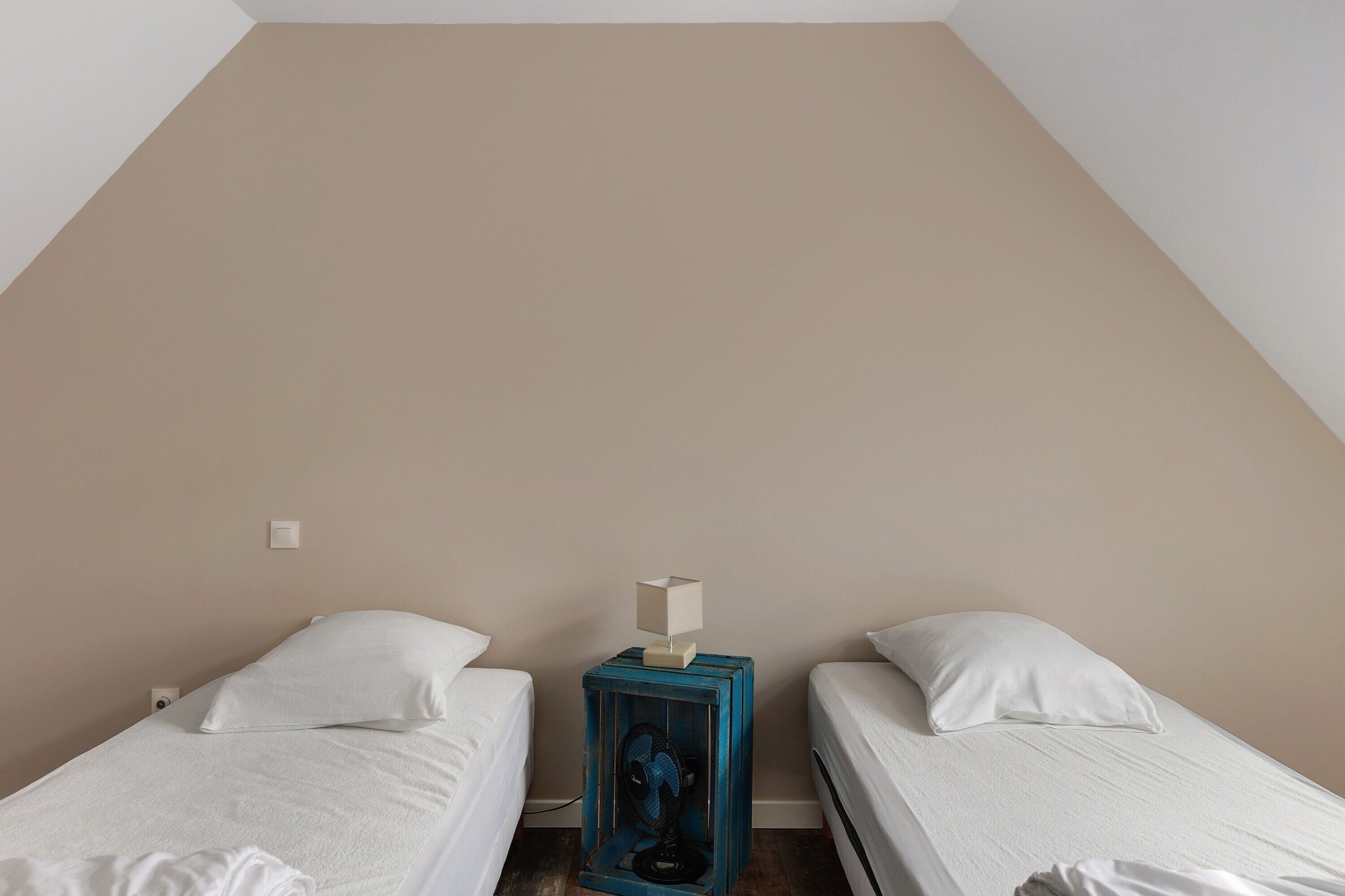 A room with two single beds, a small table with a lamp, and a wall-mounted light switch.