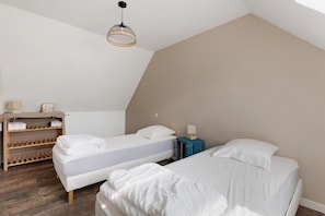 A room with two beds, a wooden shelf with bags, a hanging light fixture, and a small table with a lamp.