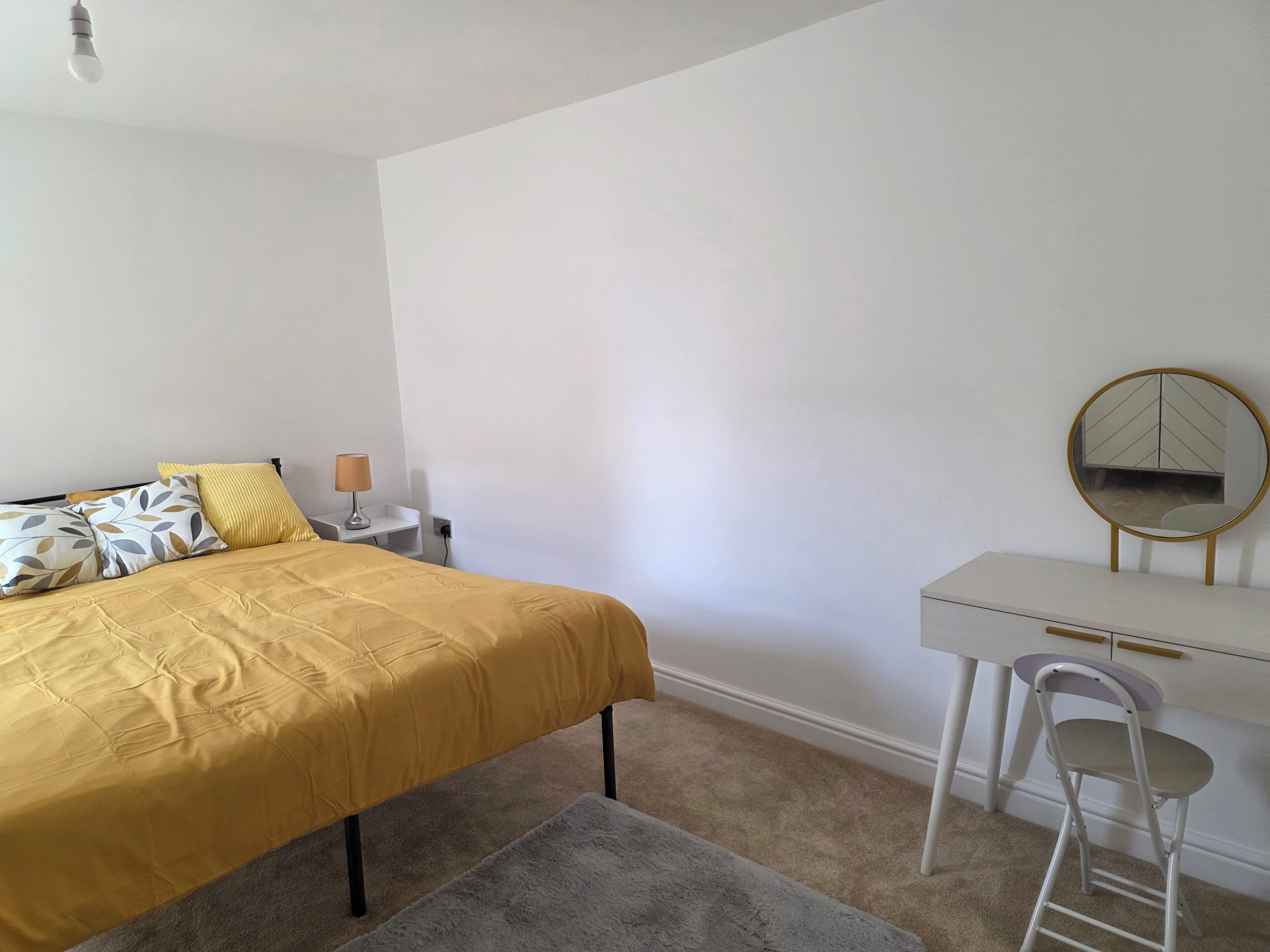 A bedroom with a bed, bedside table, lamp, desk, chair, and a round mirror.