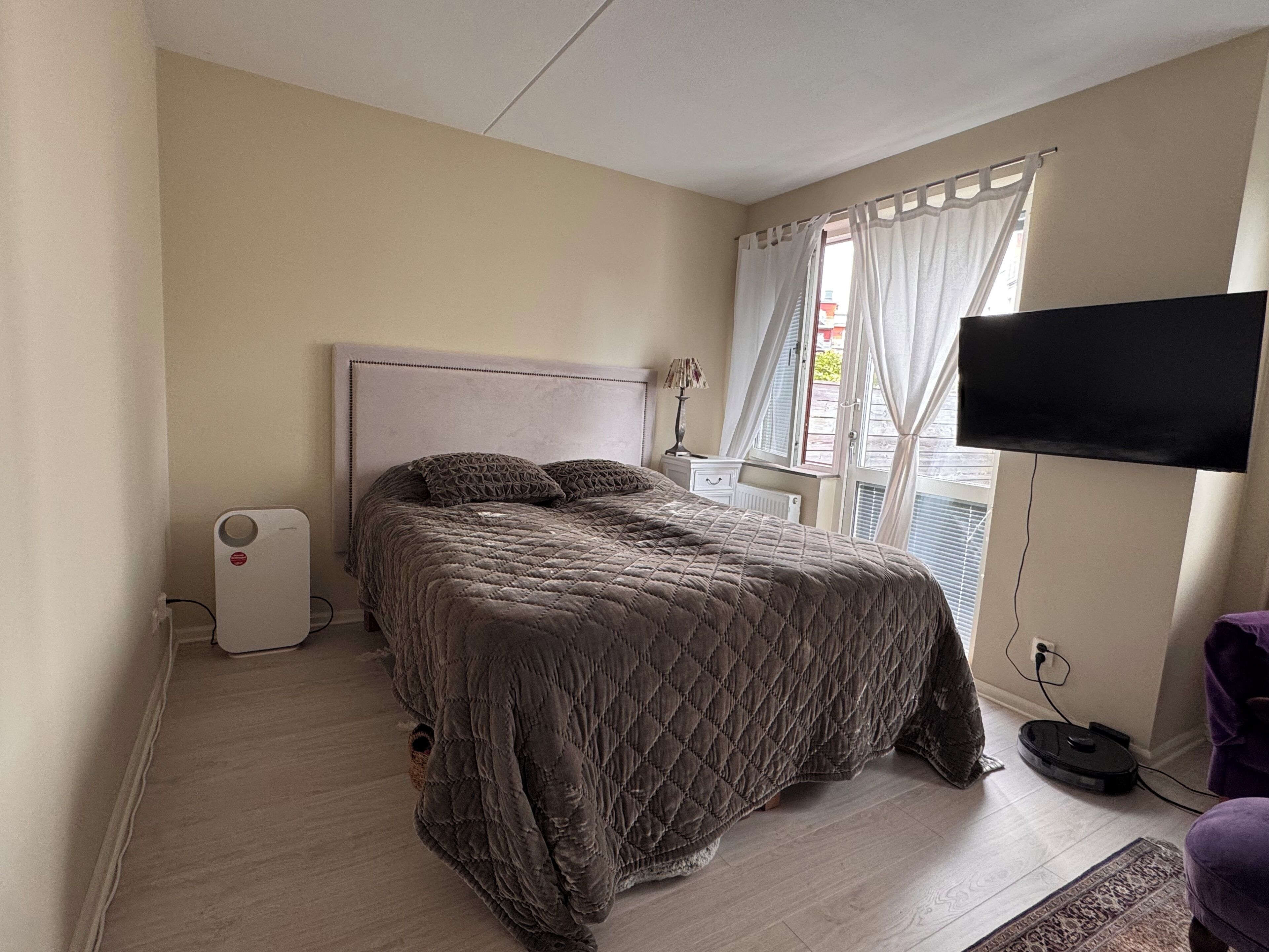 A bedroom with a bed, a television mounted on the wall, a window with curtains, and a radiator.