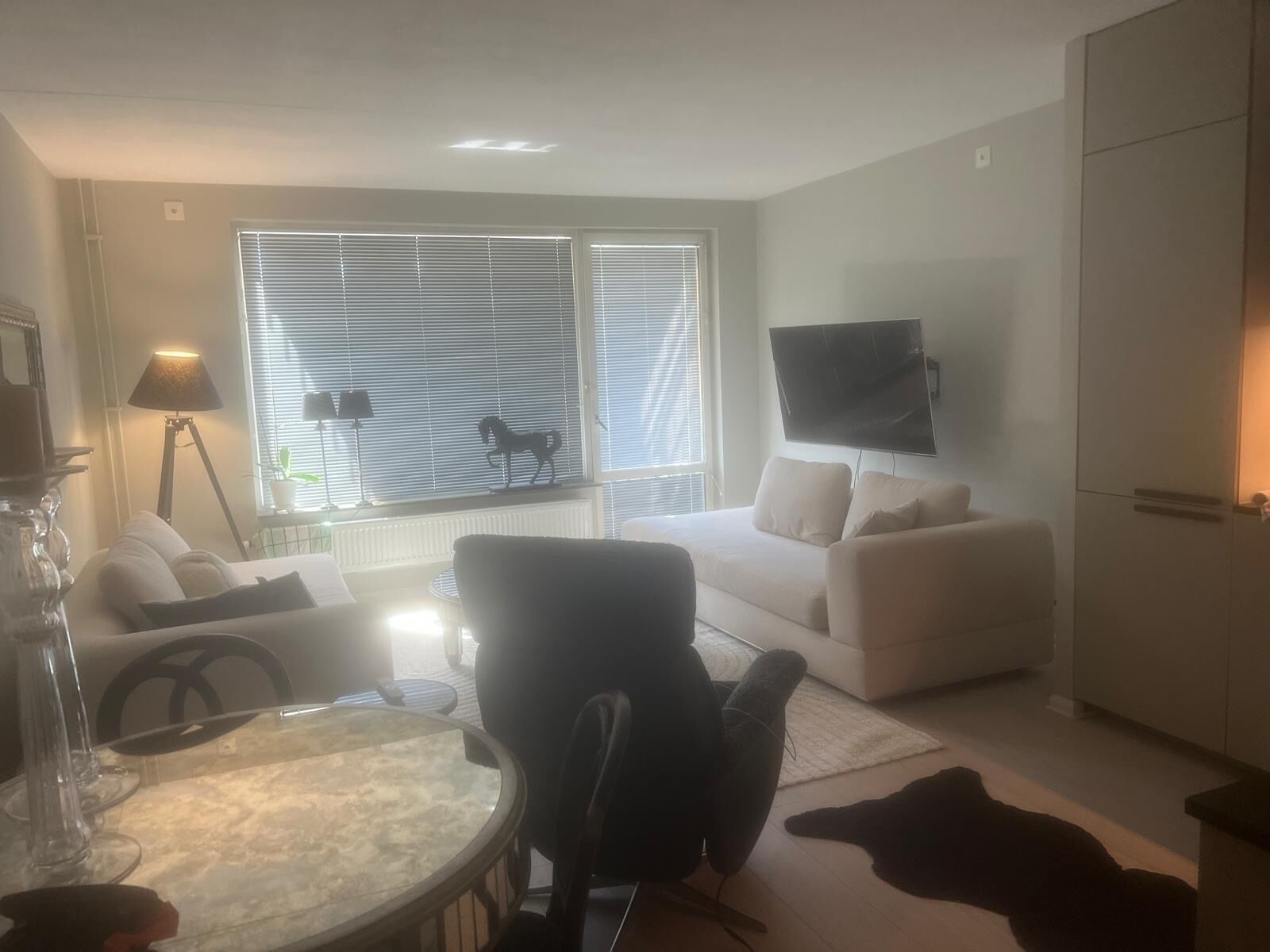A living room with a sofa, a round table, a television mounted on the wall, and a window with blinds.