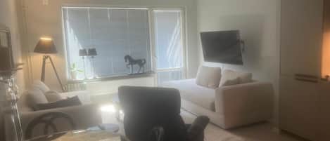 A living room with a sofa, a round table, a television mounted on the wall, and a window with blinds.