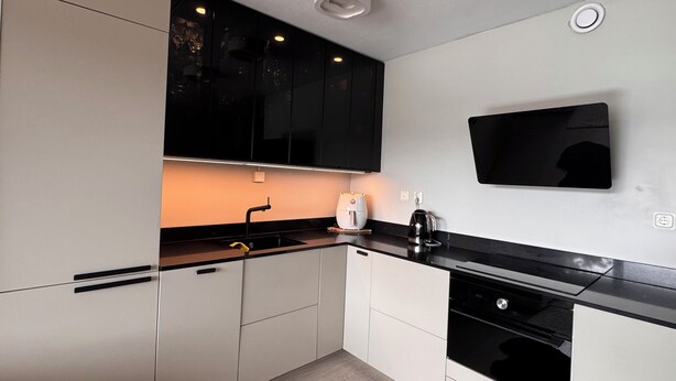 Una cocina moderna con armarios blancos y negros, encimera negra, horno empotrado y televisor montado en la pared.