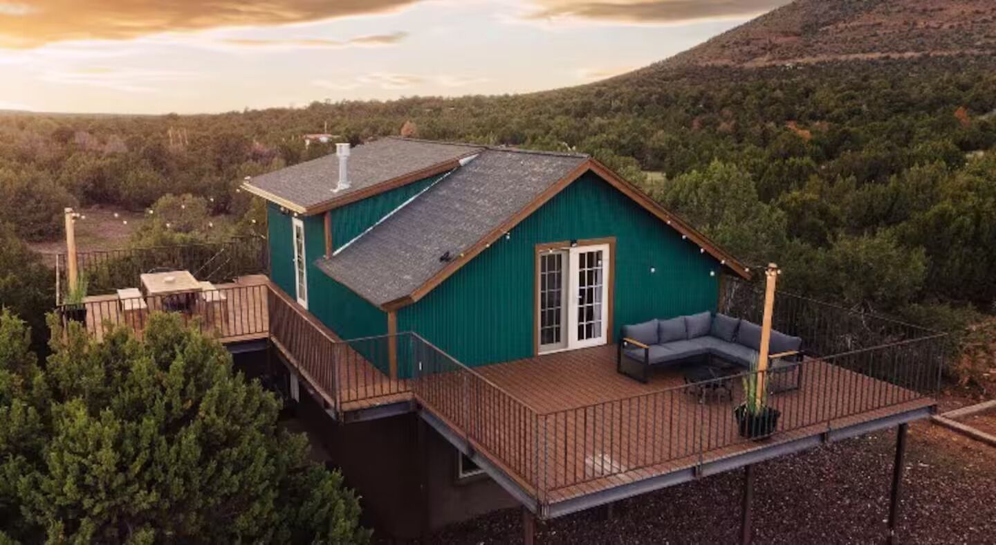 Una casa de un piso con un porche, rodeada de un bosque frondoso y un cielo despejado.
