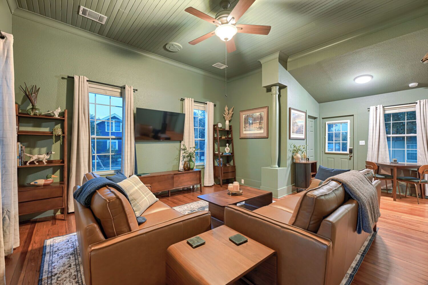 A spacious living room with a high ceiling, a ceiling fan, a flat-screen TV, leather sofas, a wooden coffee table, and a dining area with a table and chairs.