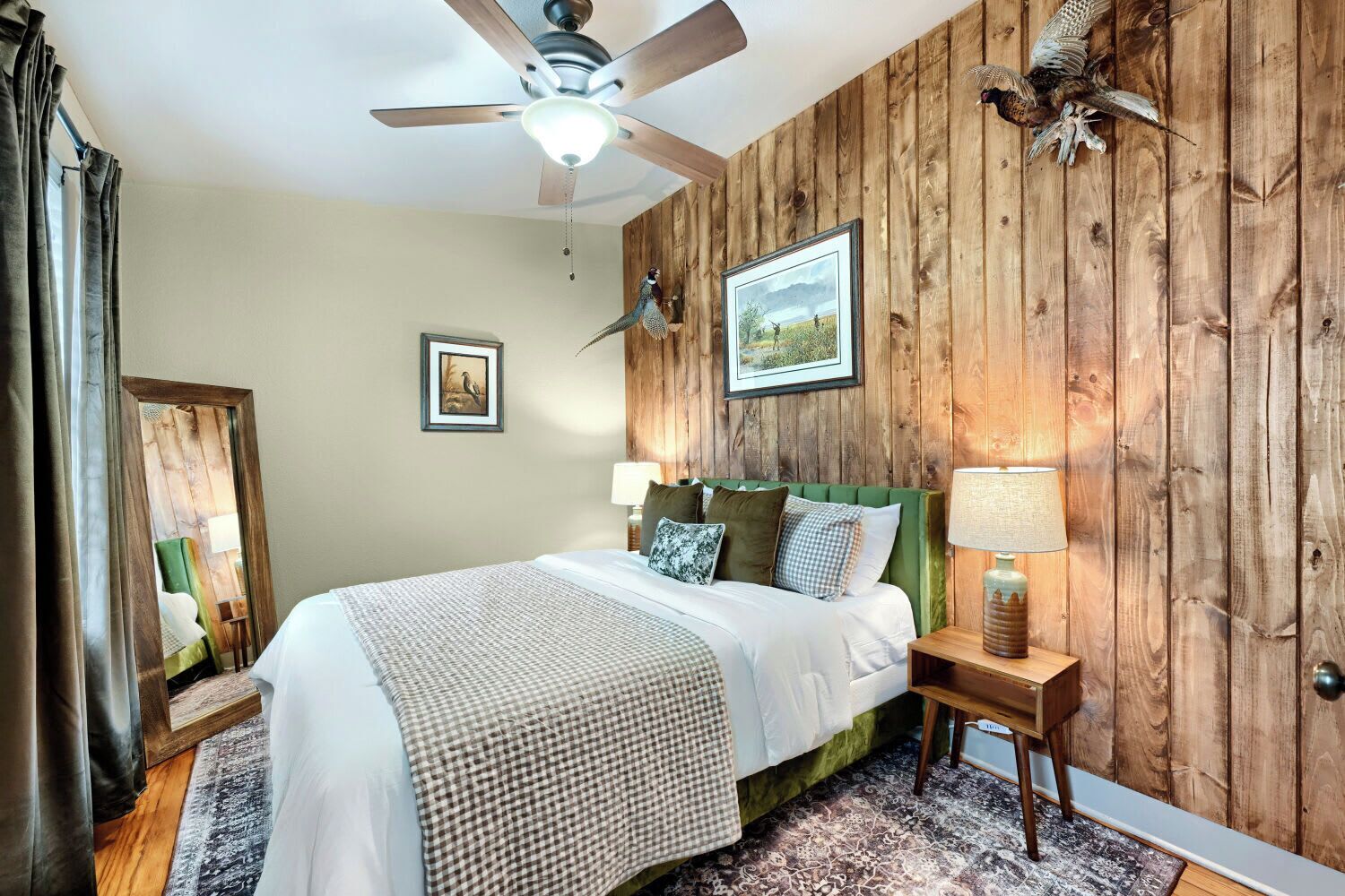 Un dormitorio con cama, ventilador de techo, paneles de madera, un cuadro enmarcado y una mesita de noche con lámpara.