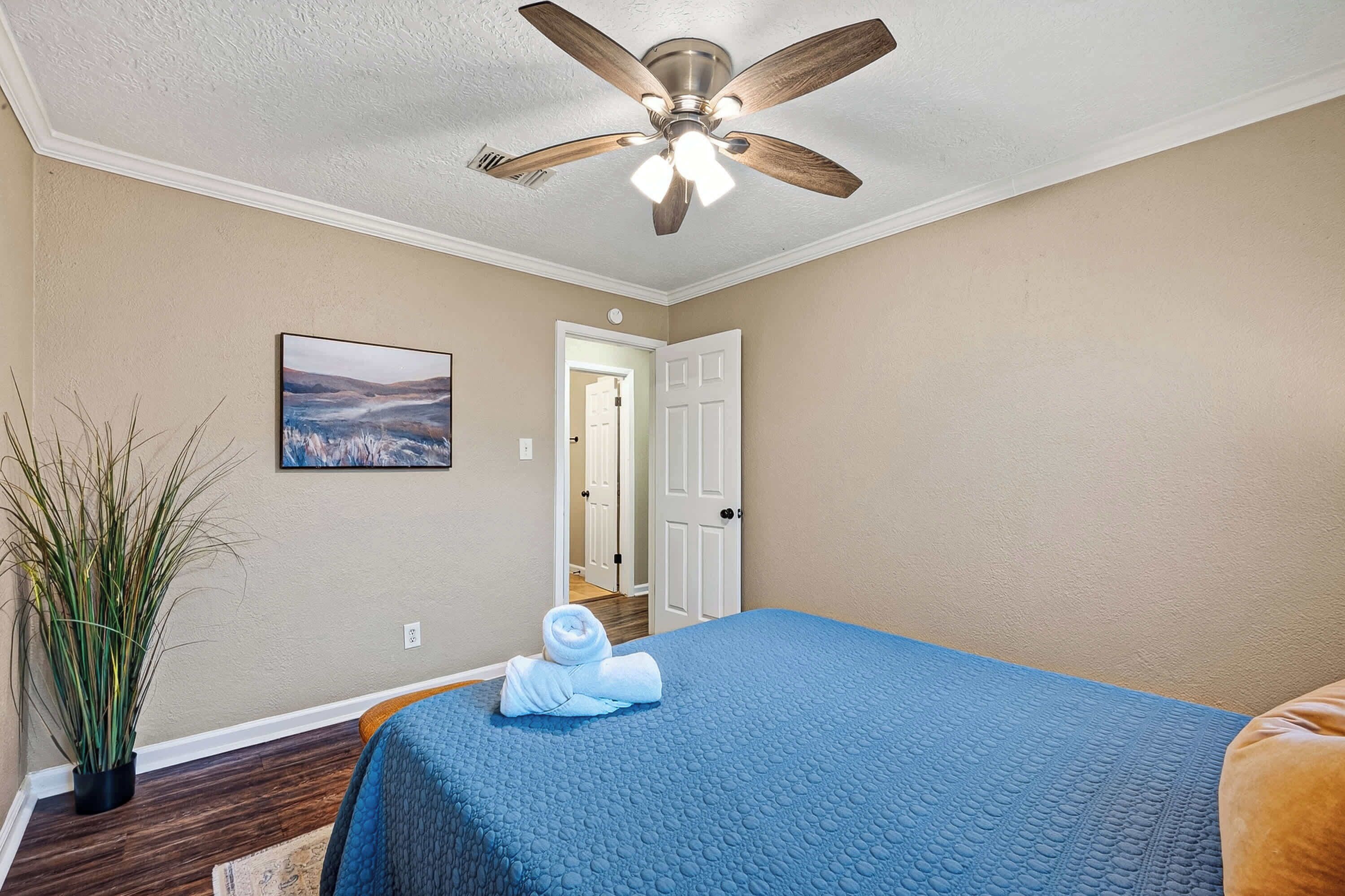 A hotel room with a bed, a ceiling fan, a door, a wall-mounted painting, and a potted plant.