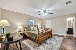 Ein Schlafzimmer mit einem Bett, einem Deckenventilator, einem Fenster mit Jalousien, einem Nachttisch mit Lampe und einem durchgehenden Türspalt erkennbaren Badezimmer.
