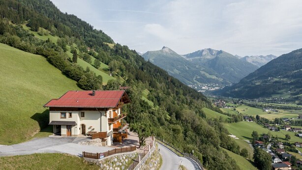 A two-story chalet with a red roof, situated on a hillside with a view of a valley and mountains in the background. - Appartements Sonnberg - Top 1 (Bad Hofgastein)
