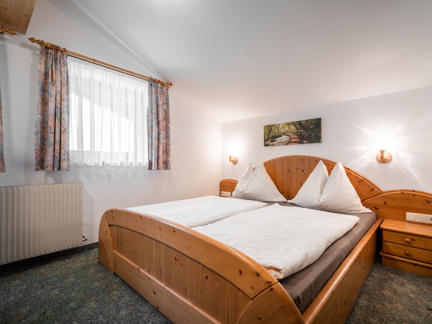 Ein Schlafzimmer mit einem hölzernen Bett, einem Heizkörper, einem Fenster mit Vorhängen, einem Nachttisch und einer an der Wand befestigten Lampe.