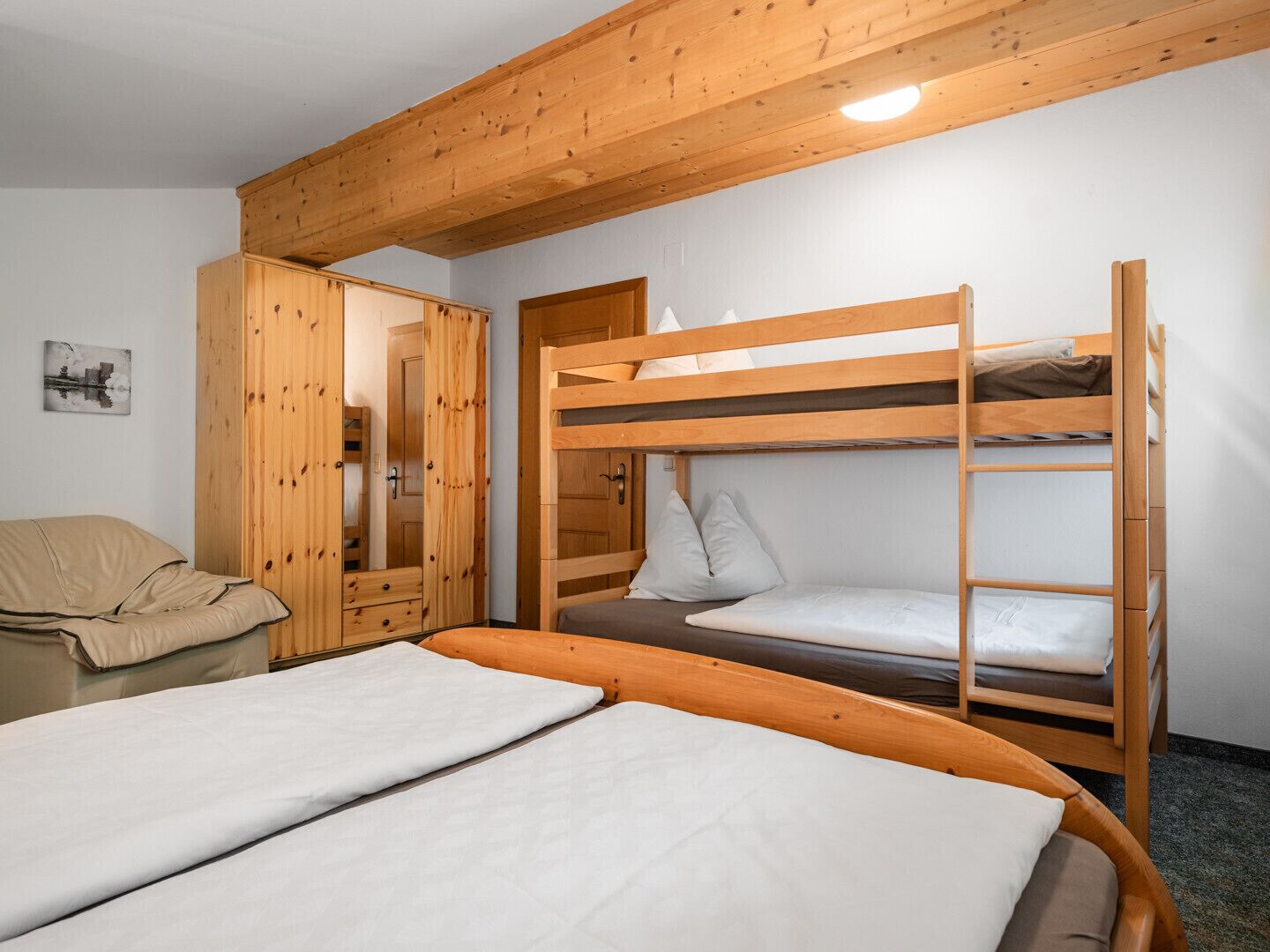 Een slaapkamer met stapelbed, houten plafond, een enkel bed met witte lakens, een houten garderobe en een stoel.