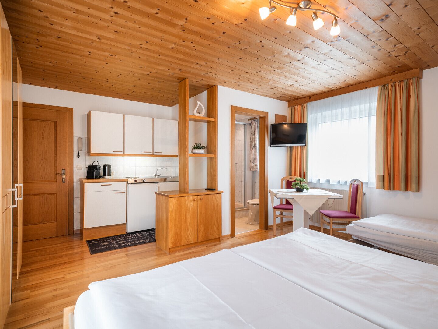 Ein Hotelzimmer mit Holzbalkendecke, einer Küchenzeile, einem Bett, einem Fernseher und einem Fenster mit Vorhängen.