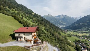 Un chalet de dos pisos con techo rojo, ubicado en una ladera con vistas a un valle y montañas al fondo.
