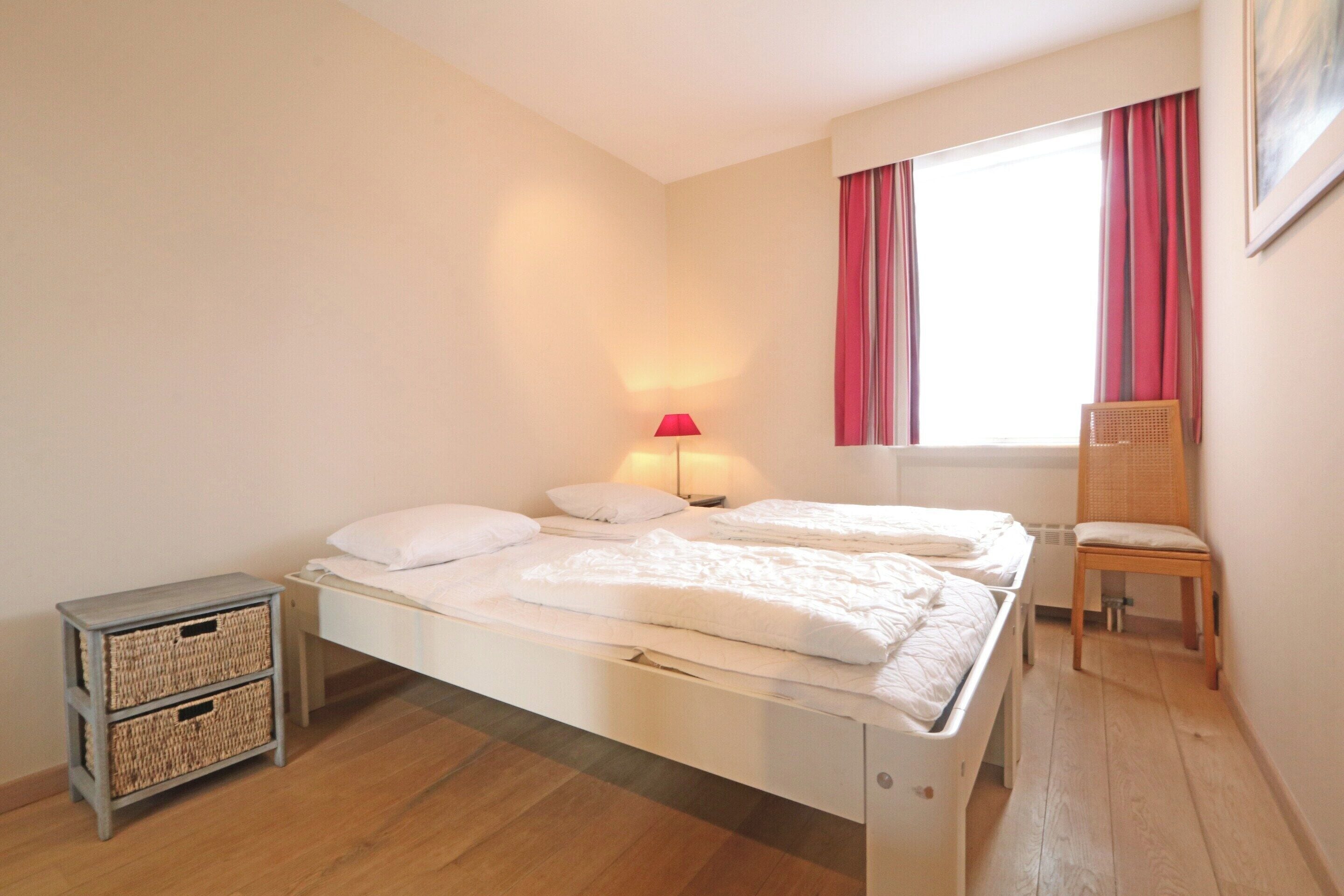 Une chambre à coucher avec un lit, une chaise, une lampe et une fenêtre avec des rideaux.