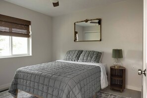 A bedroom with a bed, a bedside lamp, a mirror, and a window with blinds.