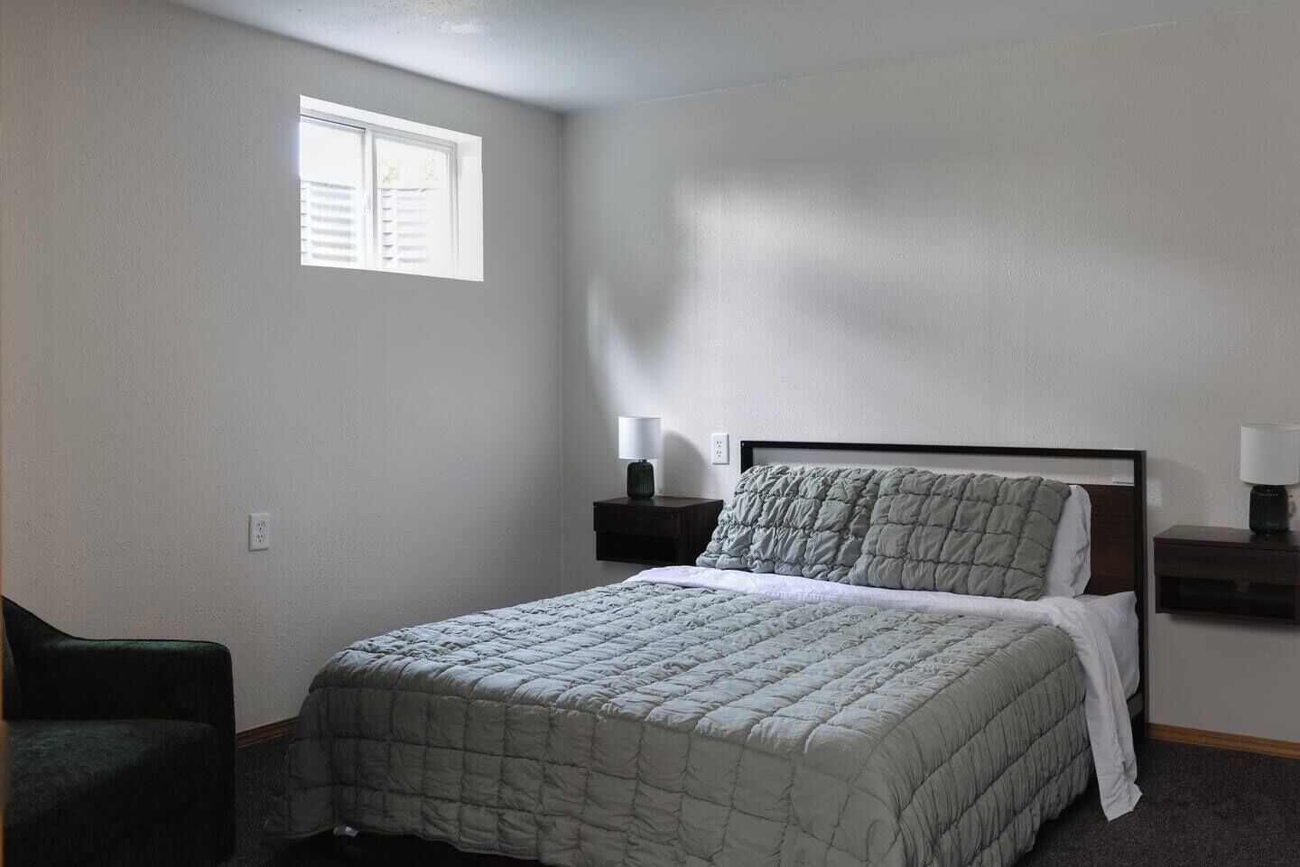 A bedroom with a bed, a nightstand, a lamp, a chair, and a window.