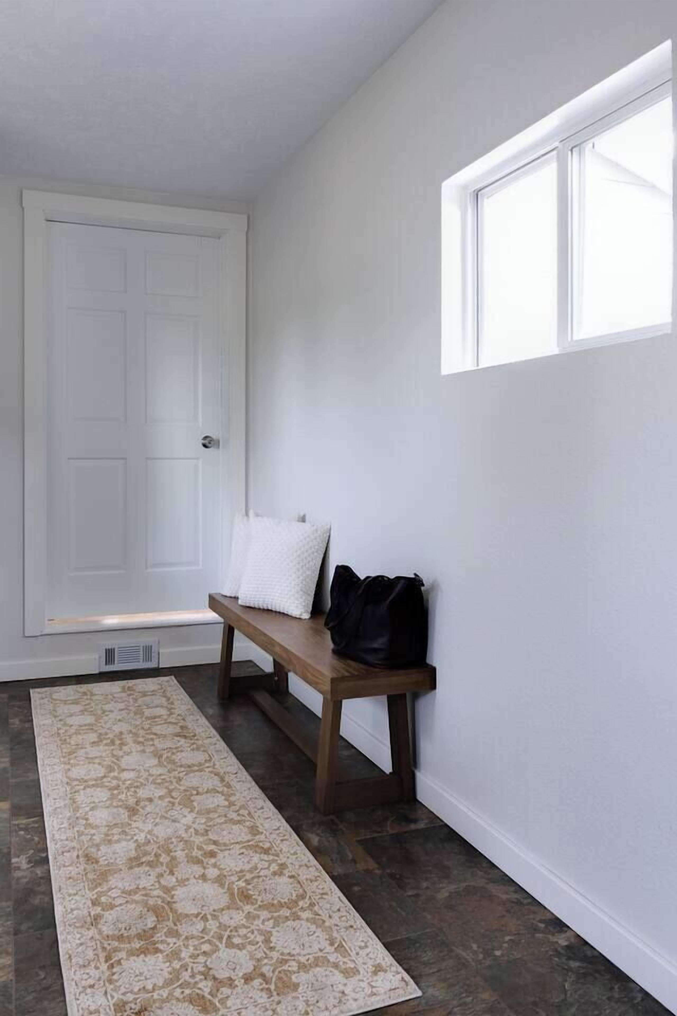 A hallway with a white door, a bench with pillows, and a patterned rug.