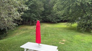 A white picnic table with a red umbrella, set on a concrete patio surrounded by grass and trees.