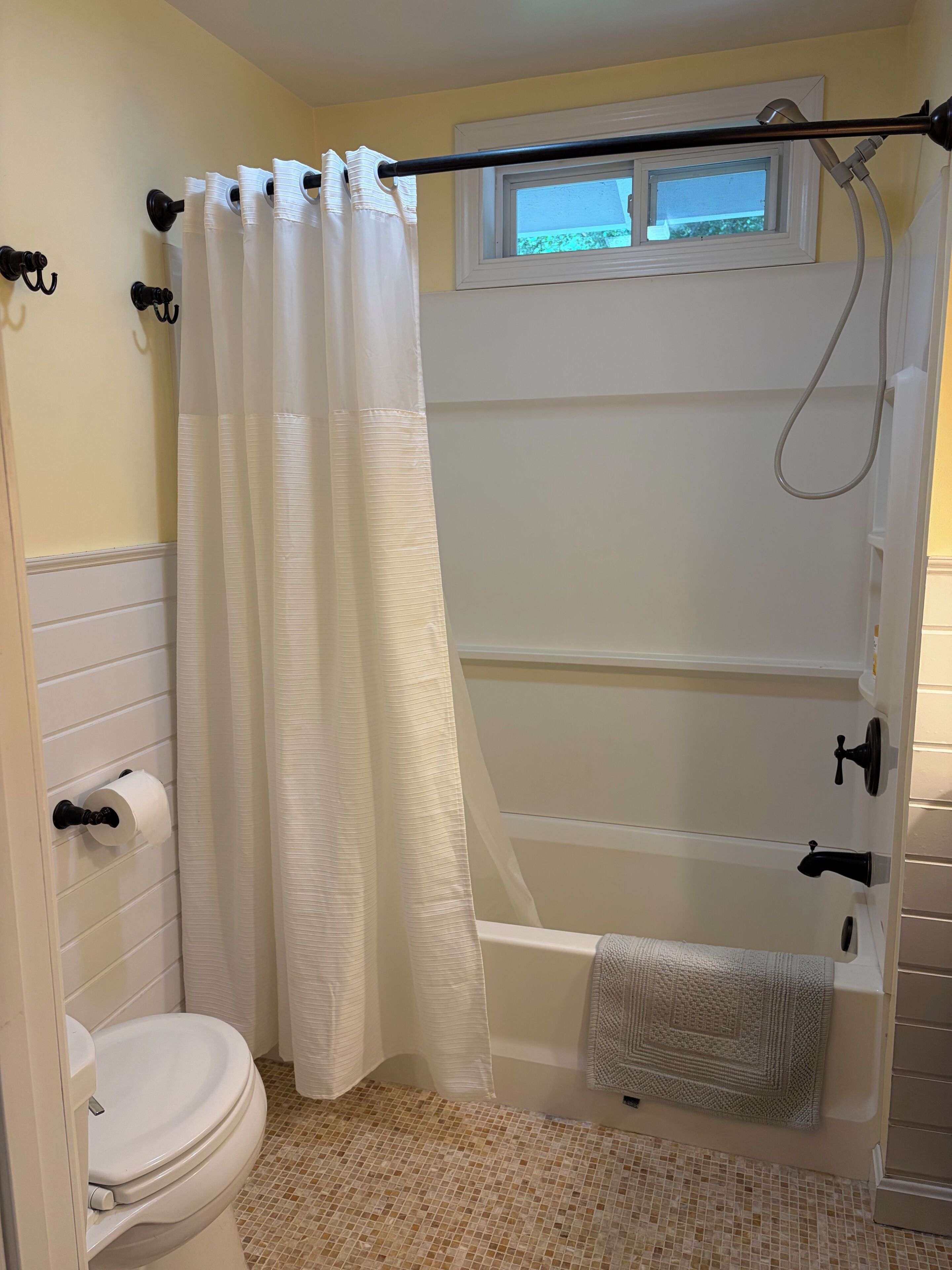 A bathroom with a toilet, a shower with a glass door, a towel rack, and a window above the shower.