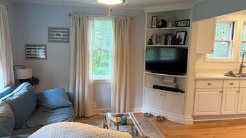 A living room with a sofa, a television, and a kitchen area with cabinets and a window.