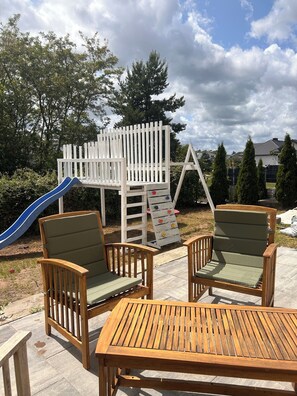 A wooden deck with two chairs and a table, a playground with a slide, and a white fence. - A spacious house with a garden by the sea, close to the beach and the pier. (Mosty)