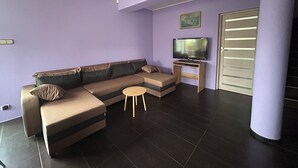 A modern living room with a sectional sofa, a small round table, a flat-screen TV on a wooden stand, and a sliding door. - A spacious house with a garden by the sea, close to the beach and the pier. (Mosty)