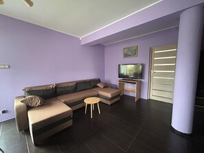 A modern living room with a sectional sofa, a small round table, a flat-screen TV on a wooden stand, and a sliding door. - A spacious house with a garden by the sea, close to the beach and the pier. (Mosty)