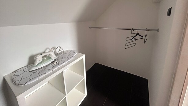 A room with a white ironing board, a hanging rack with clothes, and a dark floor. - A spacious house with a garden by the sea, close to the beach and the pier. (Mosty)