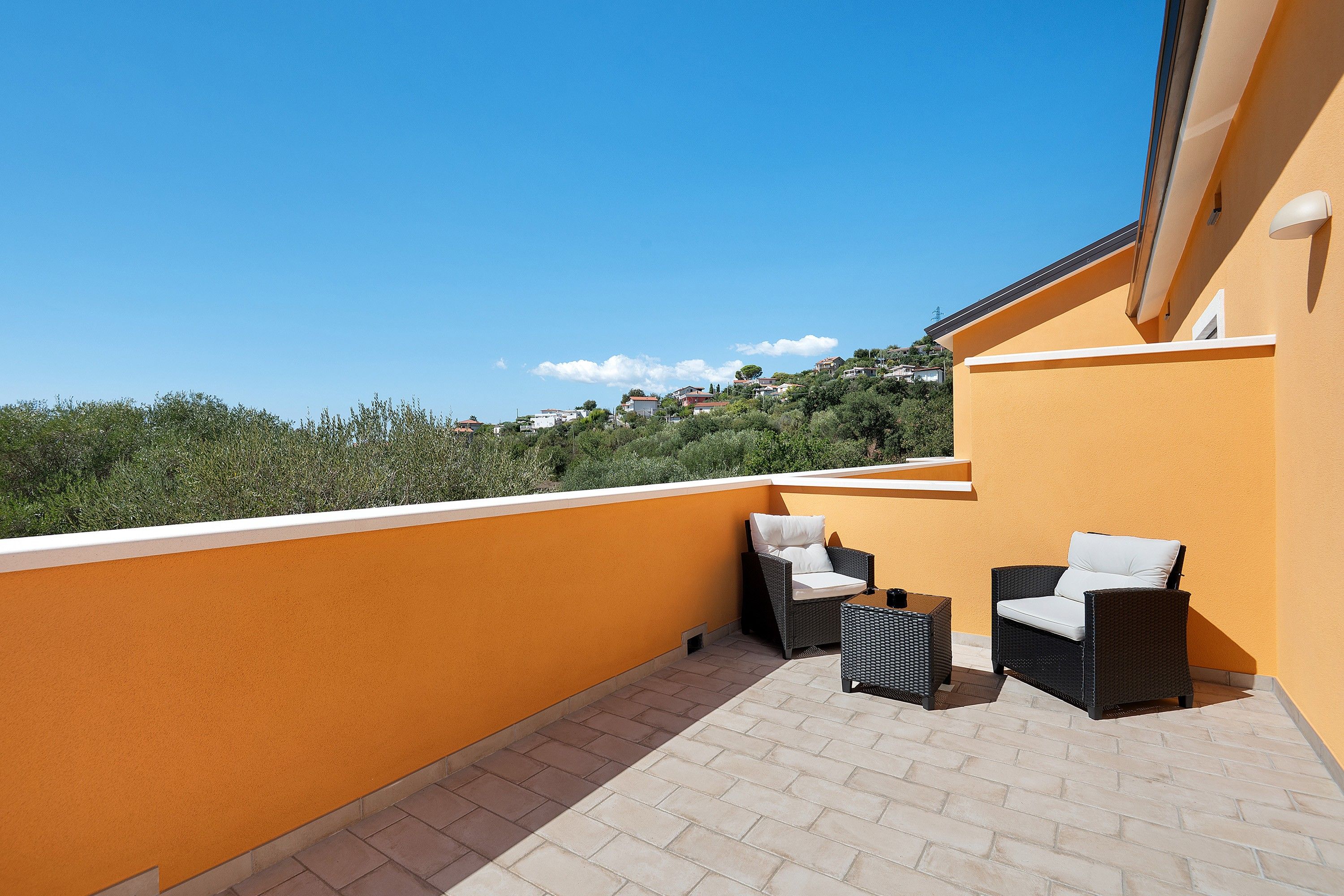 A balcony with two wicker chairs and a small table, a yellow wall, and a view of a hillside with houses.