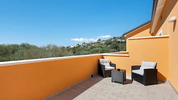 Un balcon avec deux fauteuils en osier et une petite table, un mur jaune et une vue sur une colline parsemée de maisons.