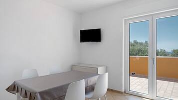 Una stanza modesta con un tavolo, sedie, una televisione a schermo piatto e una porta-finestra che si apre su uno spazio esterno.