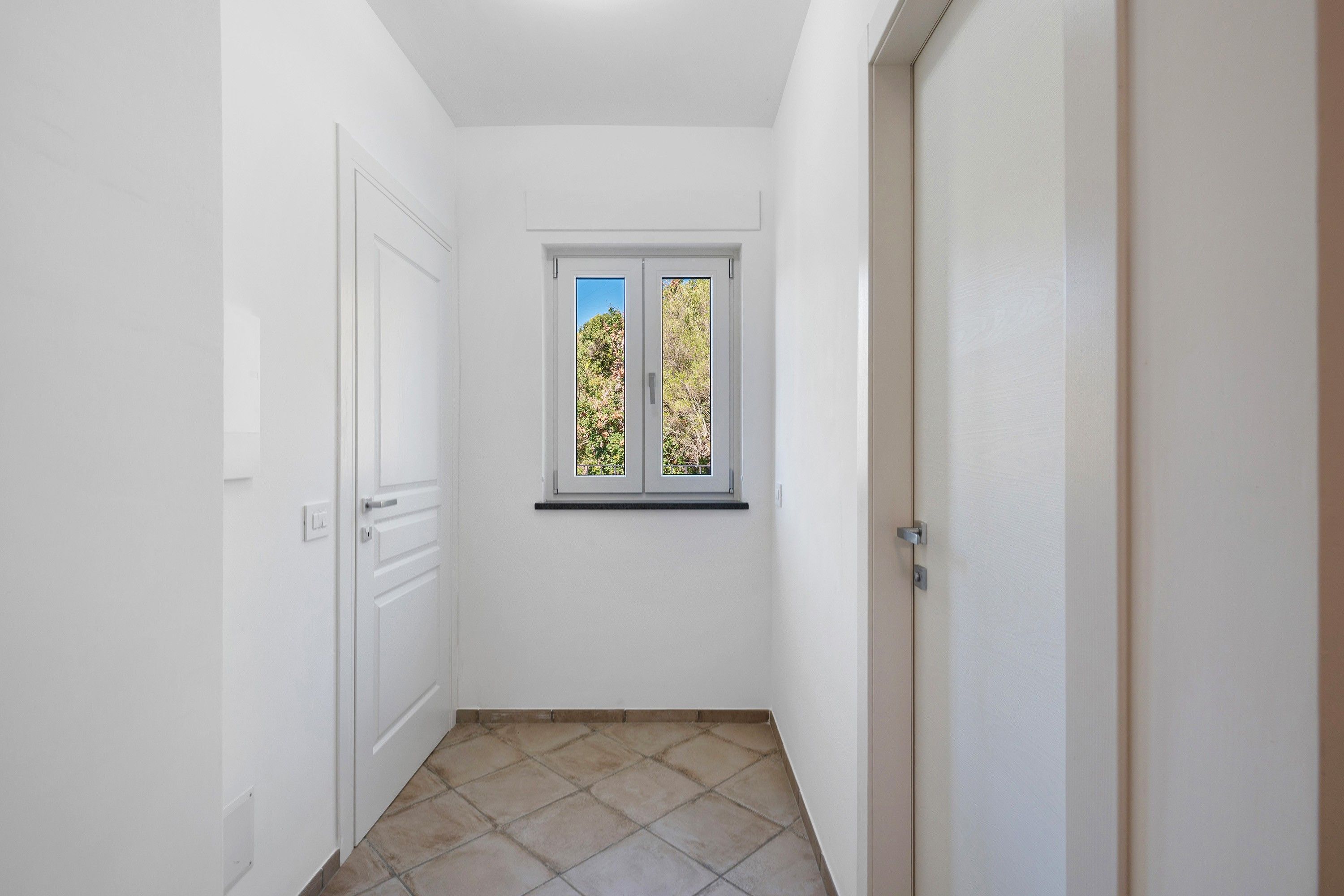 A narrow hallway with white walls, a window with a view of trees, and a door on the right.