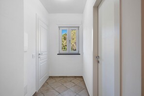 Un pasillo estrecho con paredes blancas, una ventana con vista a árboles y una puerta a la derecha.