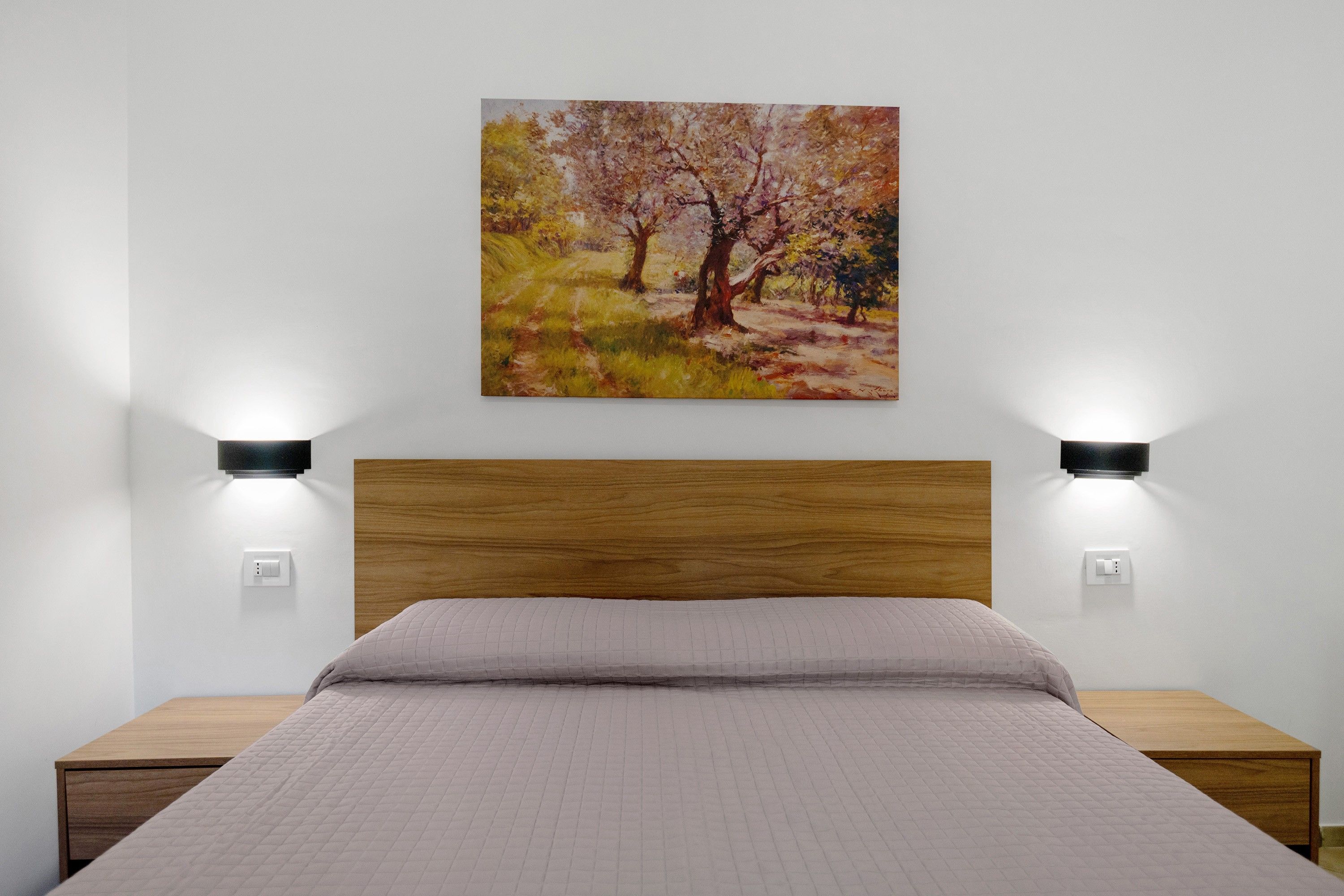 A bedroom with a wooden headboard, a bed with a quilted mattress, two bedside tables, and a wall-mounted painting.