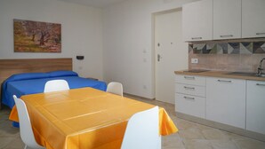 Une petite pièce comprenant un lit, une table recouverte d’une nappe jaune, un coin cuisine et un tableau accroché au mur.