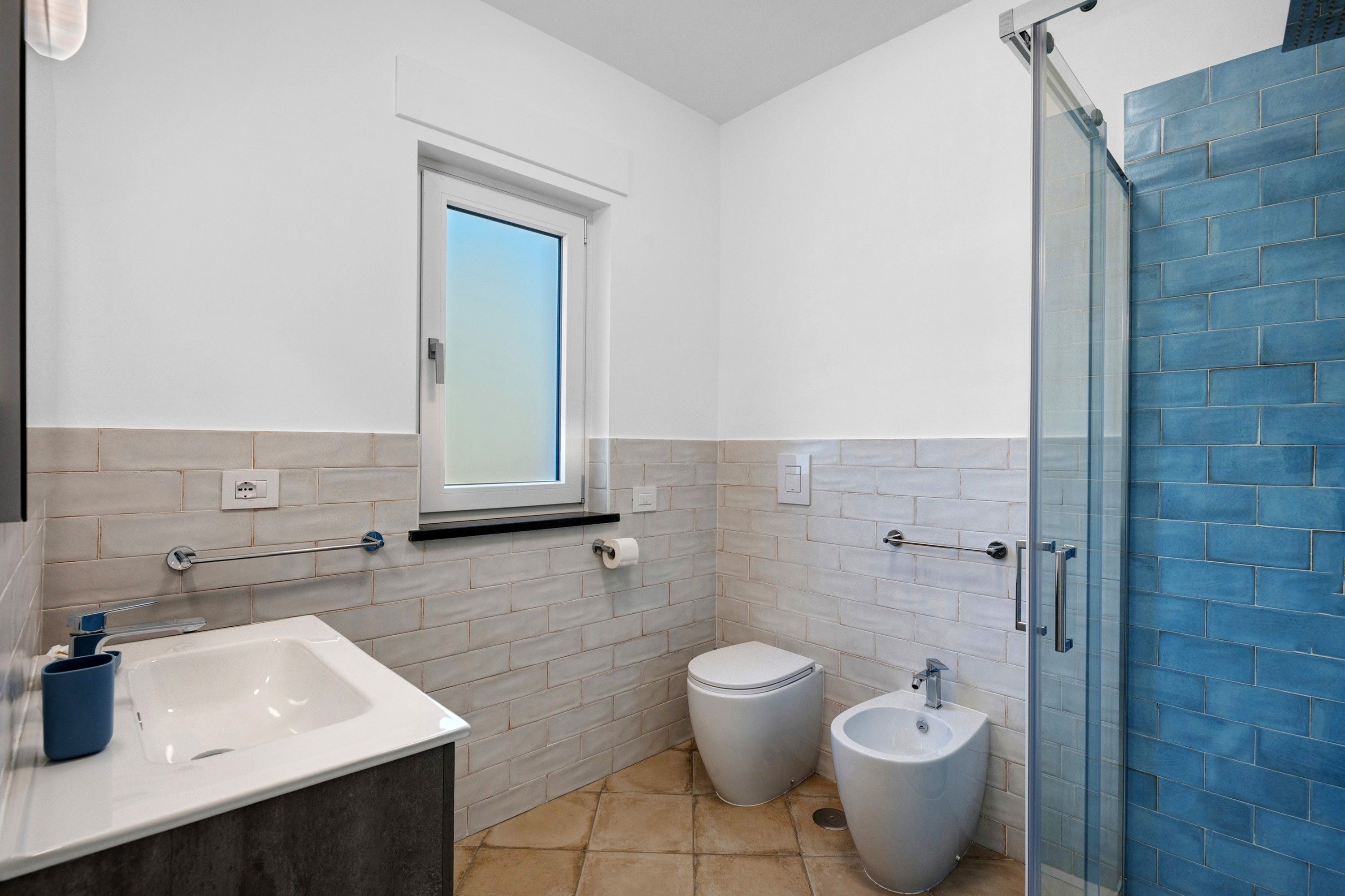 Ein modernes Badezimmer mit Waschbecken, Toilette und Bidet, einem Fenster und einer Duschkabine mit blauen Fliesen.