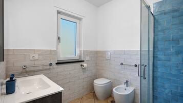 Ein modernes Badezimmer mit Waschbecken, Toilette und Bidet, einem Fenster und einer Duschkabine mit blauen Fliesen.