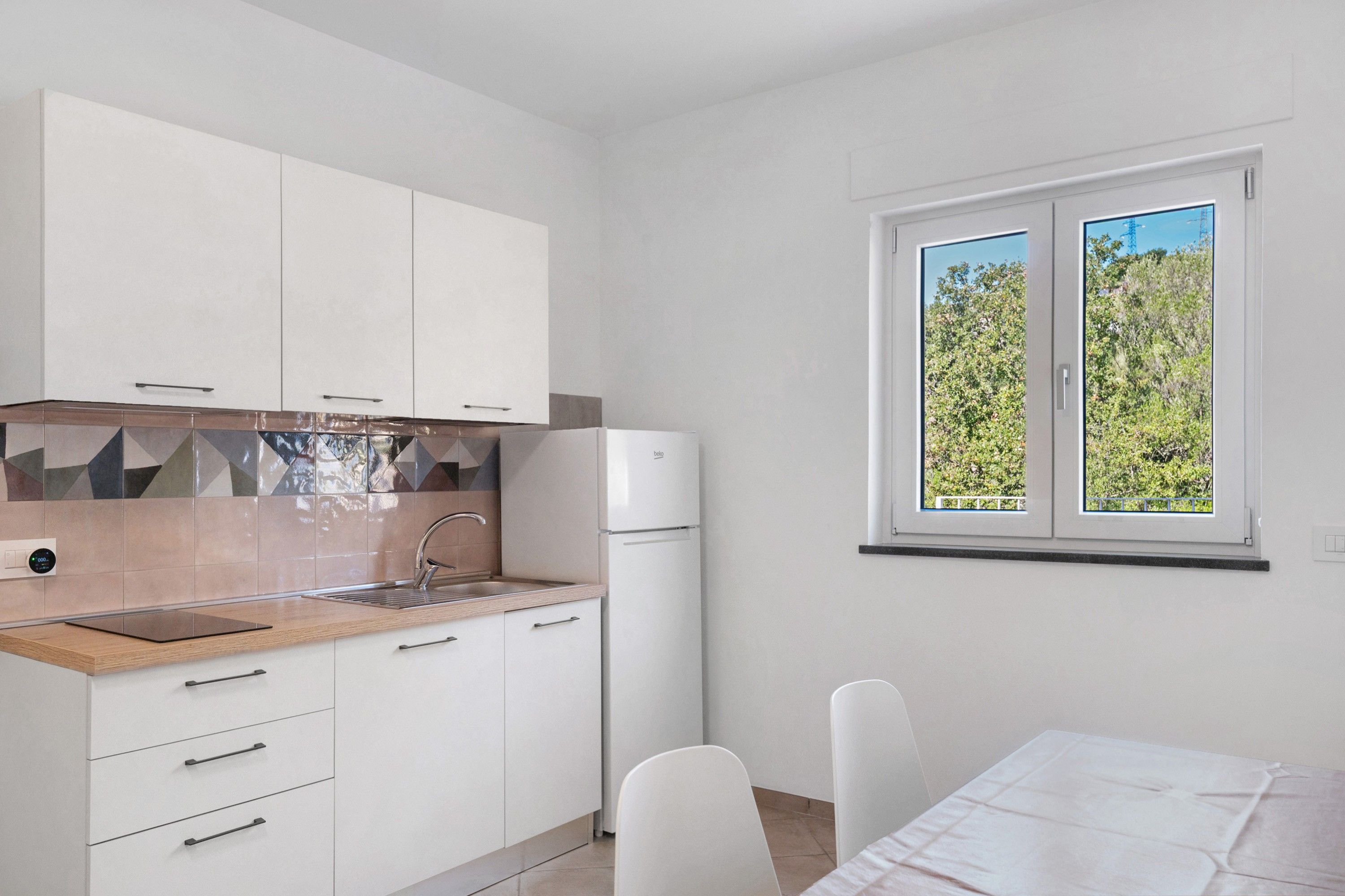 Une cuisine moderne avec des meubles blancs, un four encastré et une fenêtre donnant sur un paysage verdoyant.
