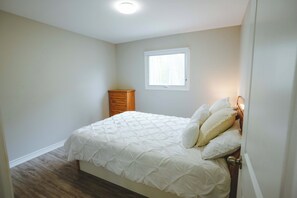 A bedroom with a bed, a window, a wooden cabinet, and a door. - Sandpiper Cottage - 3-Bedroom Cottage – Family Friendly (Trent Lakes)