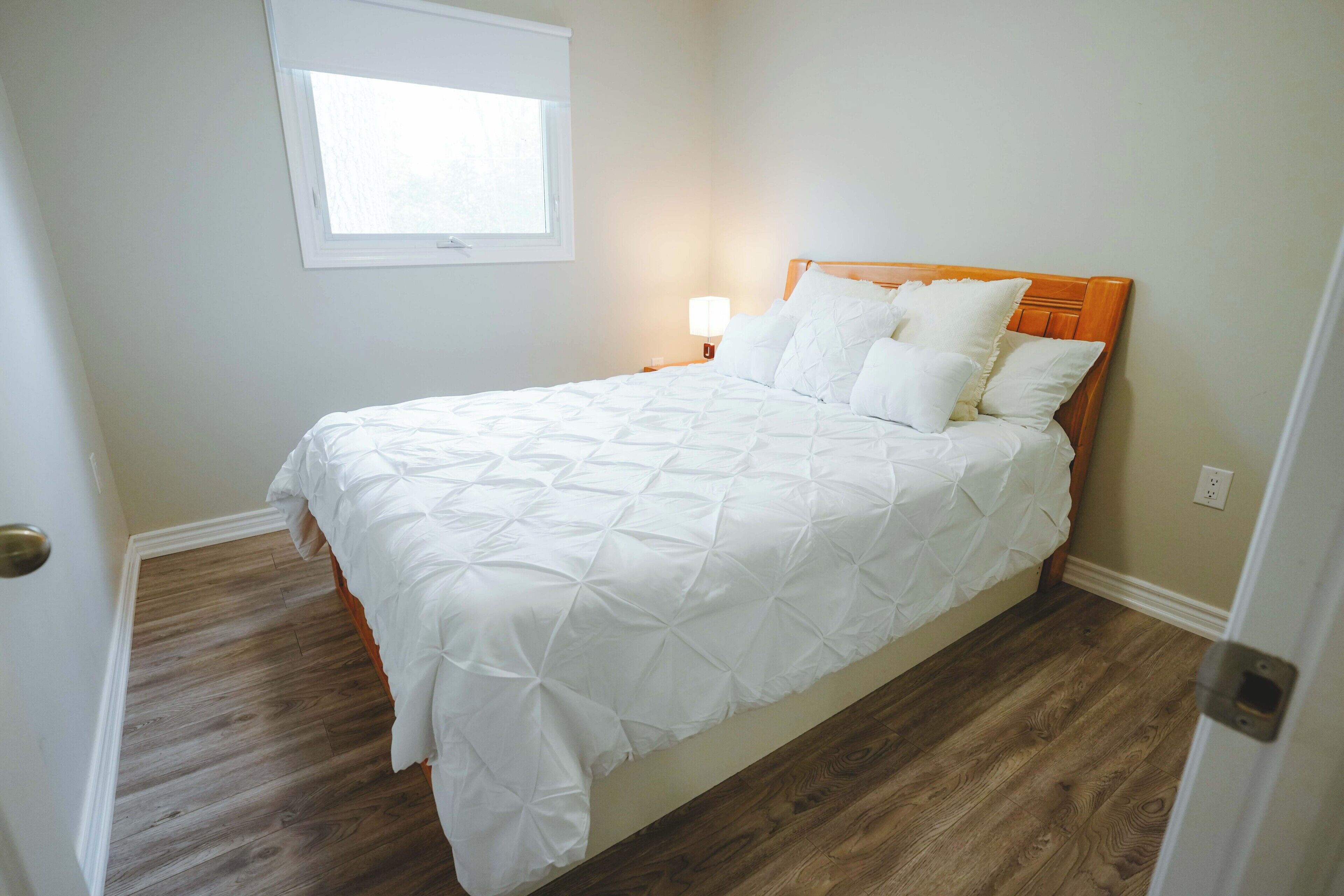 Een net opgemaakt bed met een witte deken en kussens, een houten hoofdbord, een bedlampje en een klein raam erboven.